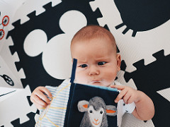 A baby sits on a black and white floor mat holding a book.