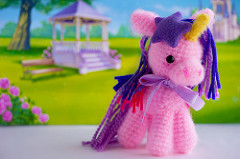 Pink crochet pony with a purple mane made of yarn stands on a table. The background is a drawing of a park.