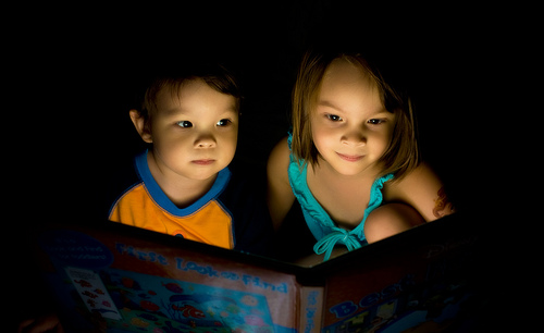 Two children sit in the dark looking at a book. The book emits light that illuminates their faces.