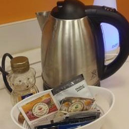 Stainless steel electric kettle with a glowing blue power light, sitting behind a small basket of assorted tea bags and snacks.