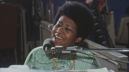 A person with a warm smile and an afro hairstyle wearing a light green shirt, leaning forward in a dimly lit indoor setting.