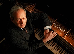 Man in a suit sits at a piano and looks up at a camera.