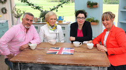 Four people sit around the table at the TV set for the Great British Baking Show.