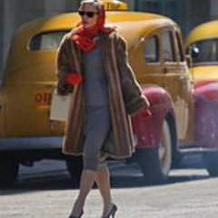 Woman with a gray dress, fur coat, and red head scarf walks down the road in front of vintage yellow taxi cabs.