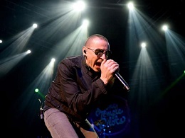 Man with close-cropped hair, sunglasses, and a black shirt on stage singing into a microphone.