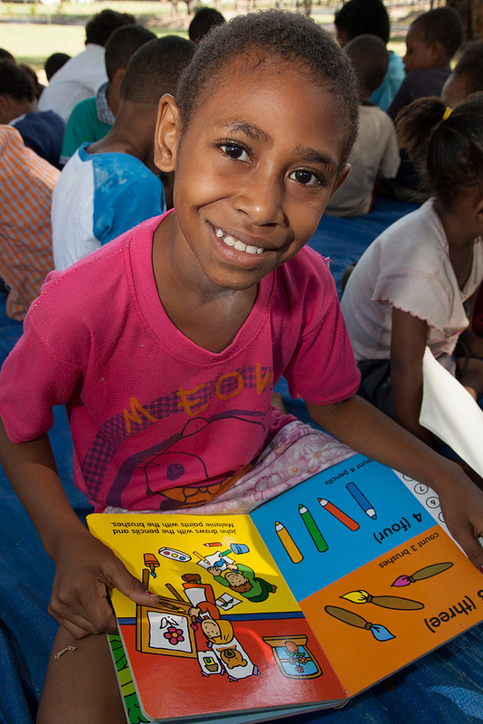 Group of children sitting on the floor. One child holds a book and smiles up at the camera.