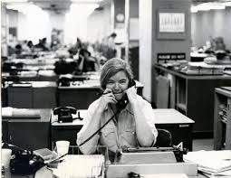 Black and white photo of a woman talking on a corded telephone at a desk in a busy, mid-century open-plan office.