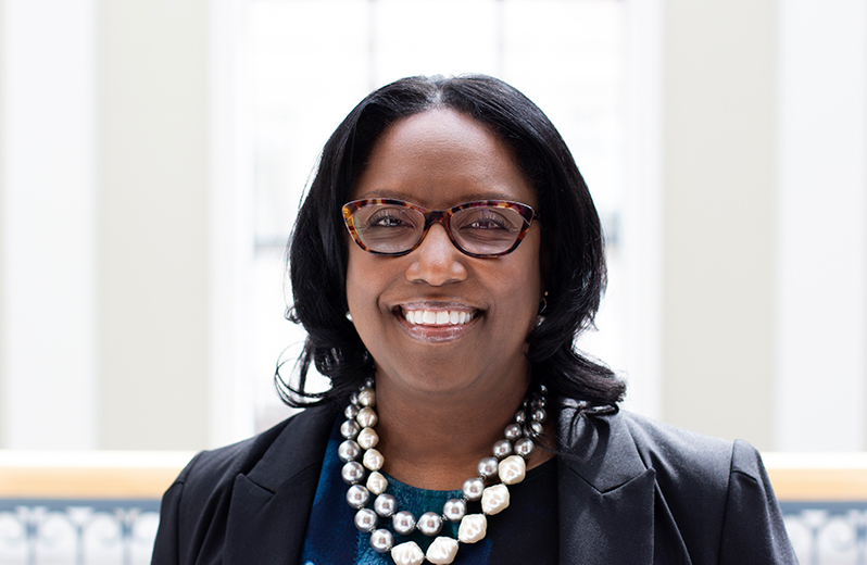 A smiling woman with glasses and a layered pearl necklace posing for a professional headshot against a bright, neutral background.