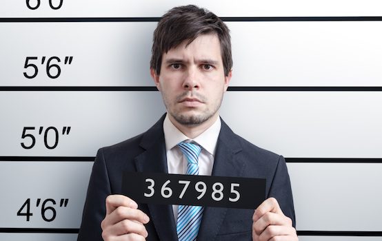 Man with dark hair and a suit and blue tie stands in front of a height chart at a police station.