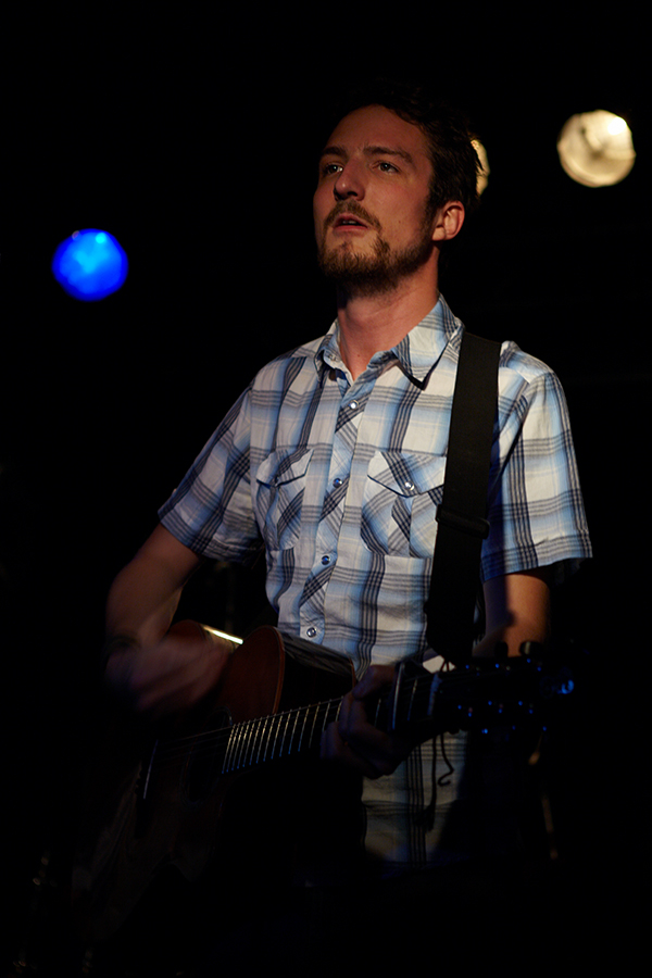 Man in a white, blue, and black plaid western shirt stands on stage playing a guitar.