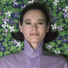 Gloria Vanderbilt in a purple blouse against a green and purple floral background.