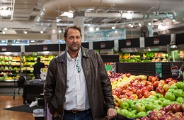 Man in a blue shirt and leather jacket stands in a grocery produce department.