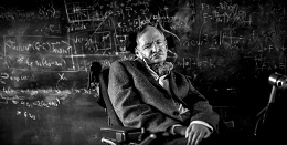 Black and white photo of Stephen Hawking in his wheelchair, positioned in front of a chalkboard covered in equations.