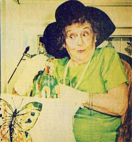 Vintage-style photo of an older woman in a green dress and large black hat, looking surprised next to a butterfly.