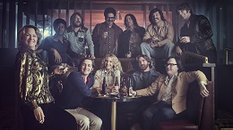 A group of people in 1970s attire pose in a dimly lit bar with drinks, featuring various hairstyles and vintage clothing.