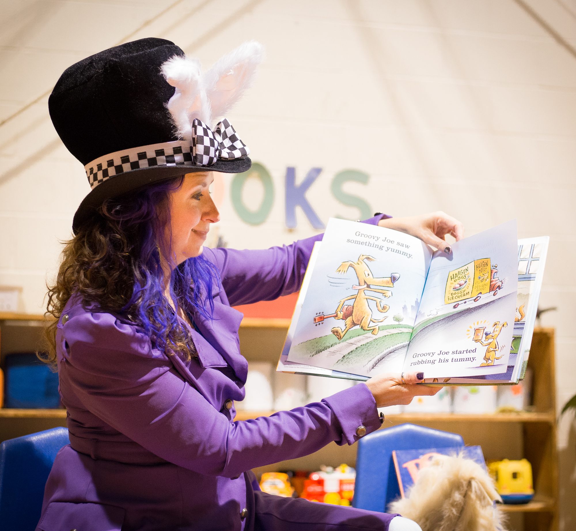 Woman with long curly brown hair wears a black top hat and bunny ears and a purple shirt while holding a children's book and reading to a crowd.