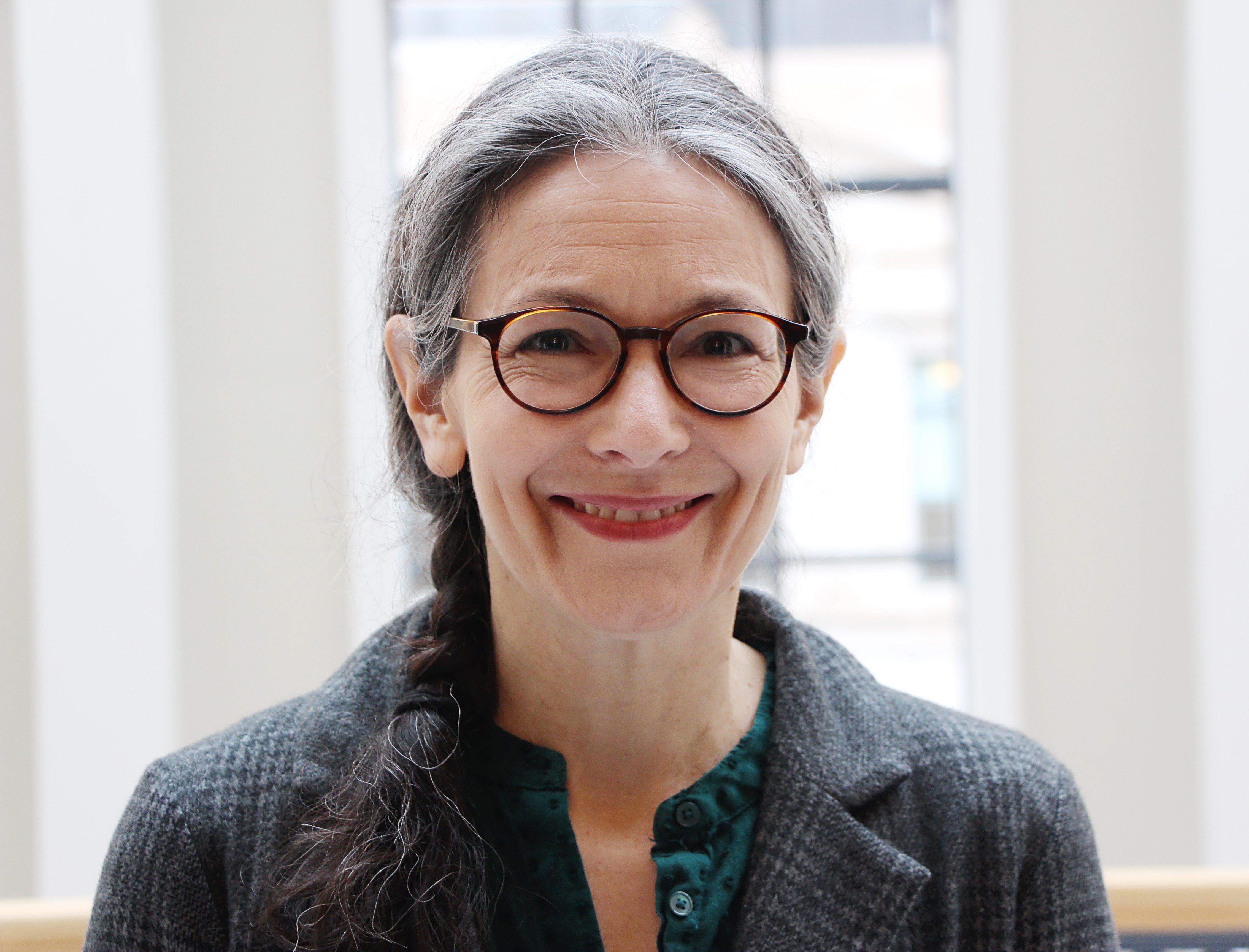 Headshot of a woman with a gray shirt, dark glasses, and long gray hair pulled back, standing in a well-lit foyer.