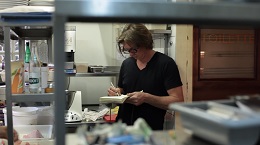 Jeff Martin in a black shirt preparing food in a commercial kitchen, seen through a service window.