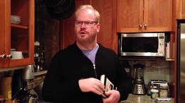 A man with reddish hair and glasses wearing a black sweater over a light purple shirt, standing in a kitchen with wooden cabinets.