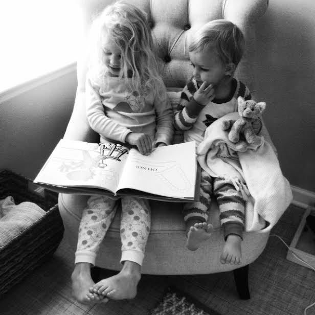 Black and white photo of two young children sitting together in a large armchair, looking at an open book.