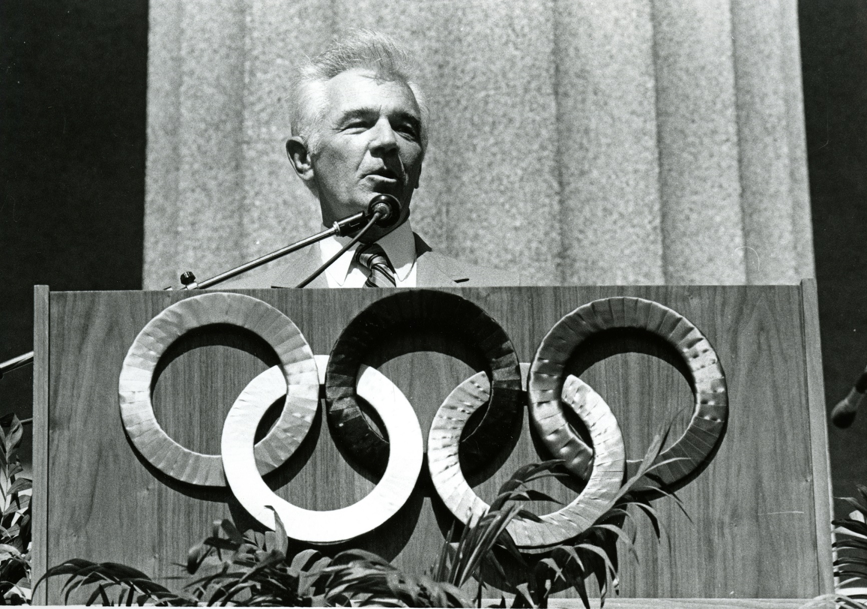 Black-and-white photo of a man speaking from a podium decorated with the Olympic rings against a fluted backdrop.