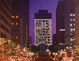 Crowded street scene shows a skyscraper lit with a message, arts music dance.