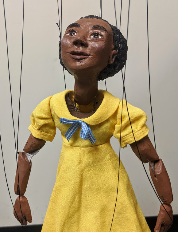 Marionette depicts black girl wearing yellow dress.