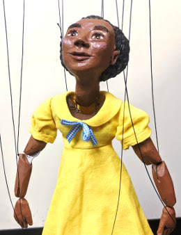 Marionette depicts black girl wearing yellow dress.