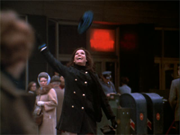 woman in dark overcoat tosses a red beret in the air.
