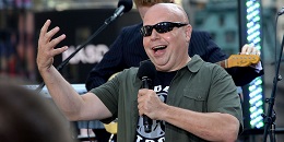 Seated man gestures with arm, holding microphone.