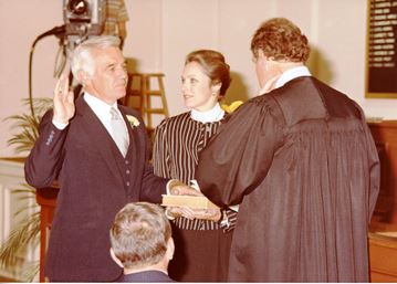 Judge in robes holds out book, while man holds hand on it, and holds right hand aloft.