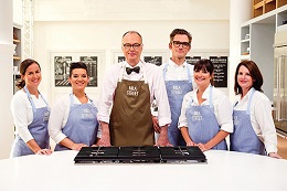 six people wearing purple aprons stand at a kitchen counter