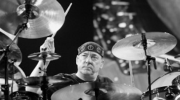 Black and white photo of Rush drummer Neil Peart wearing his signature kufi cap while playing a large drum kit.