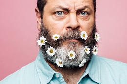 Nick Offerman with daises in his beard against a pink background.