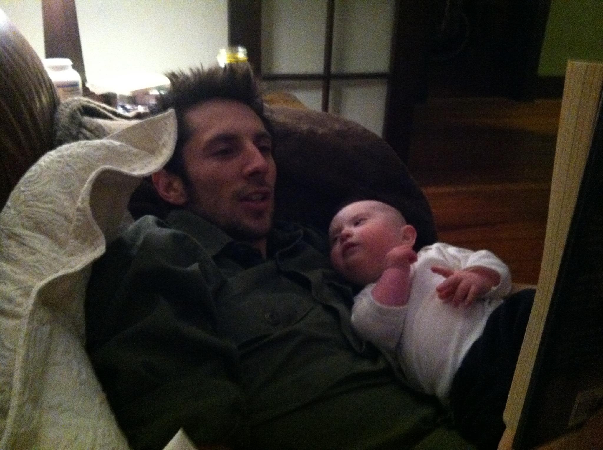 A man relaxes on a couch, holding a baby on his chest while reading a book aloud in a dimly lit room.