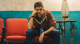 A man with a beard sits pensively on an orange chair against a teal wall, resting his chin on his hand in a dim room.
