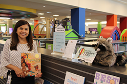 Woman with long brown hair and a white blouse holds a children's book next to a bookcase at a library.