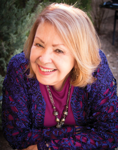 A smiling woman with blonde hair wearing a purple patterned cardigan and a magenta shirt, looking toward the camera.