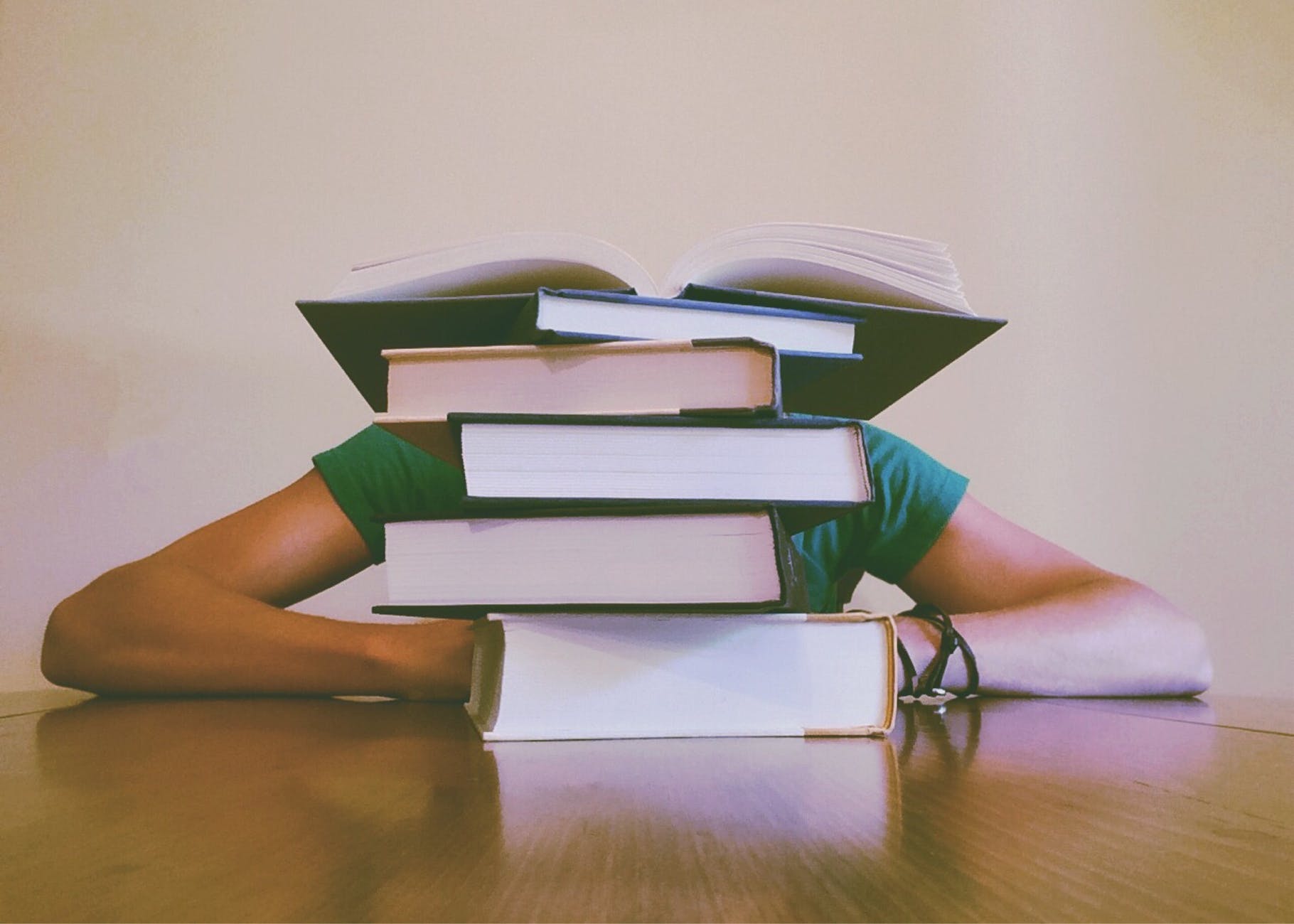 Man sits at a table with his head and torso hidden by a pile of books.