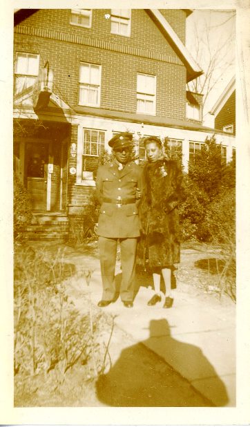 Vintage sepia photo of a soldier in uniform standing with a woman in a fur coat in front of a brick house.