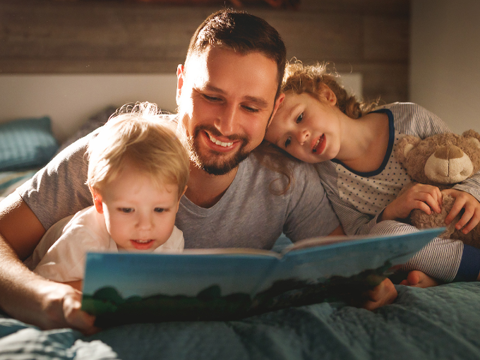 Father laying on a bed with his son and daughter reading a book.