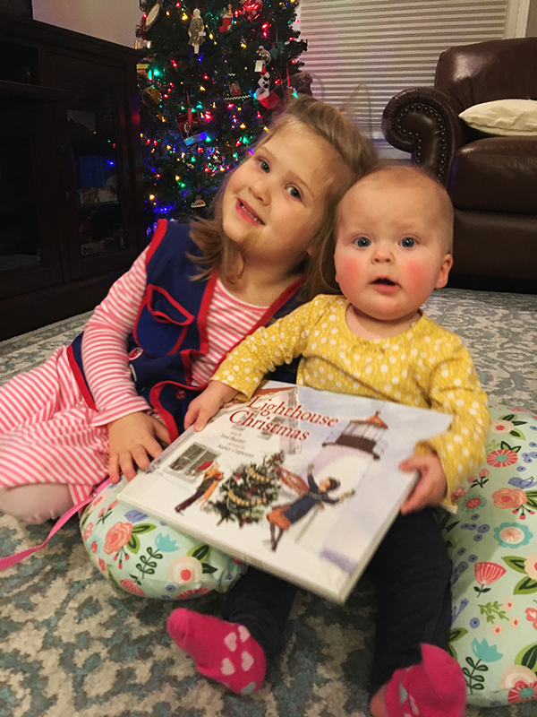 A young girl and a baby sit on a patterned rug holding a Christmas book, with a lit holiday tree in the background.