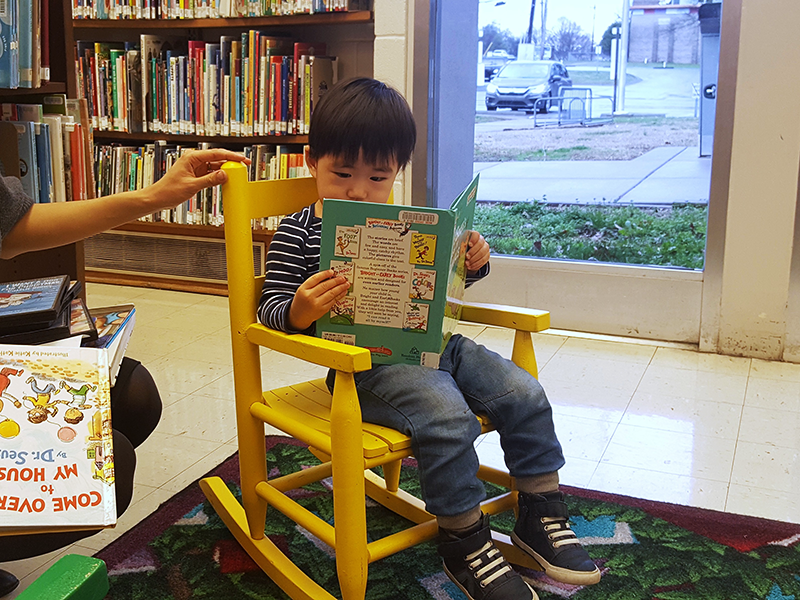 Young boy with dark hair sits in a child-sized yellow rocker and reads.