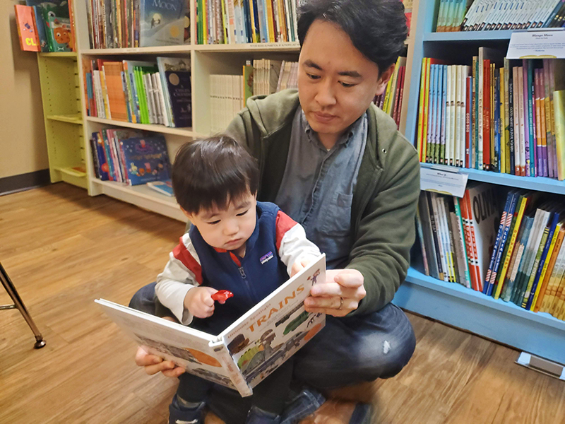 Young boy with father sits on the floor in front of bookcases and reads a book.