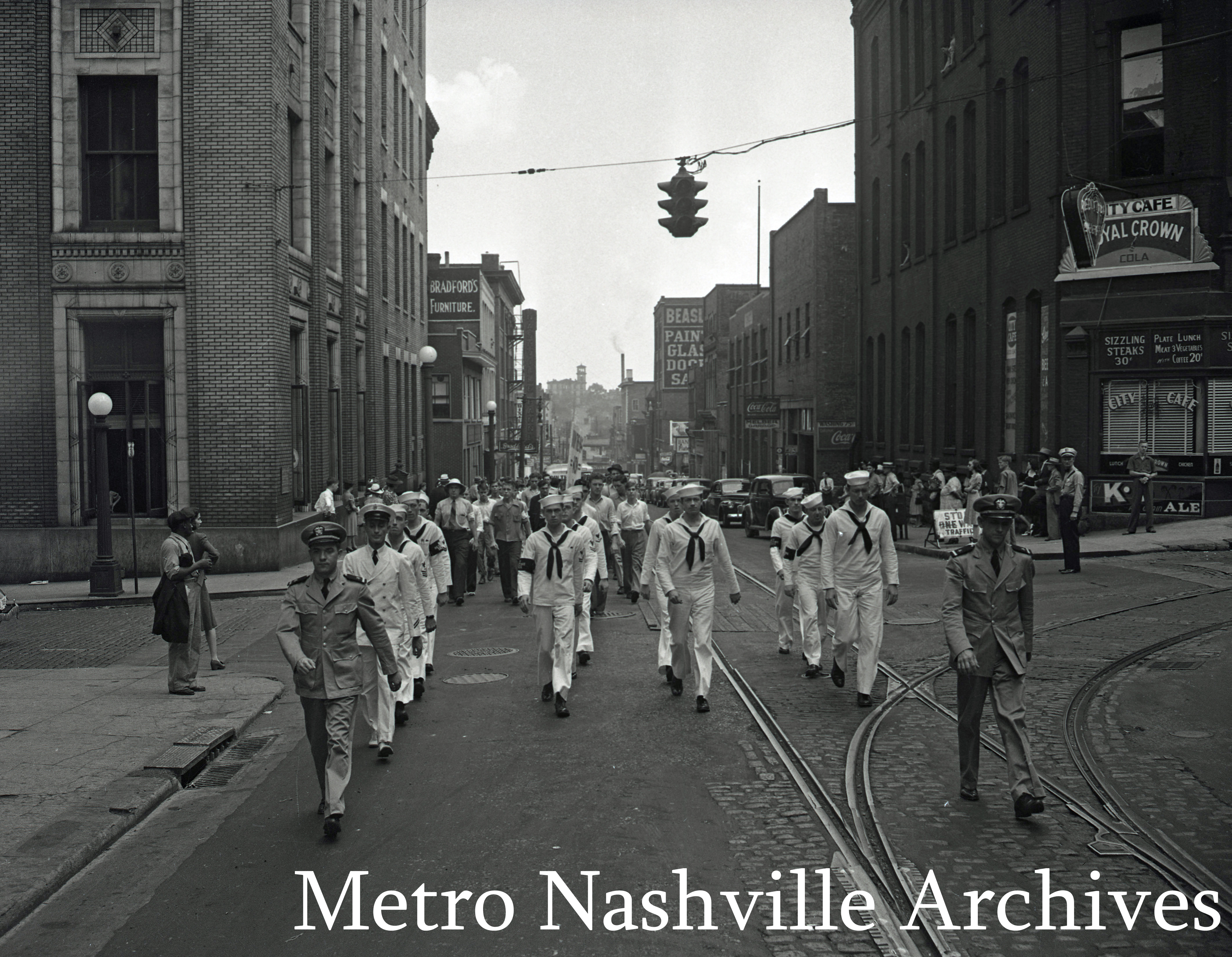 Men in white sailor uniforms march in line down city street.