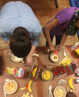 Overhead shot of two women plating bananas over oatmeal on a long table.