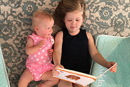 A young girl in a black tank top reads a picture book to a baby in a pink floral outfit.