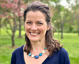 Smiling brown-haired woman in blue shirt and necklace, standing in park.