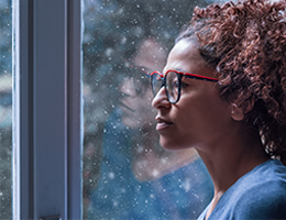 Profile of Black woman in glasses by window, looking melancholy.