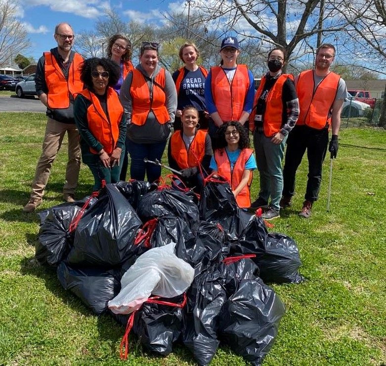 Group of orange-vested people on lawn standing in front of pile of full trash bags. 